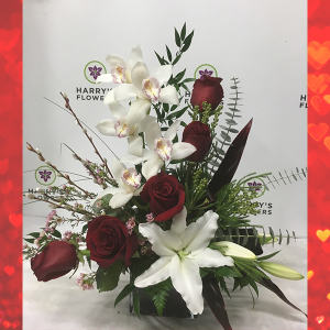 red roses, cymbidium orchids, white oriental lilies, green berzelia, pink wax flowers, stems of pussy willow, red cordyline leaves, eucalyptus and mixed greenery all in a deep burgundy red glass cube